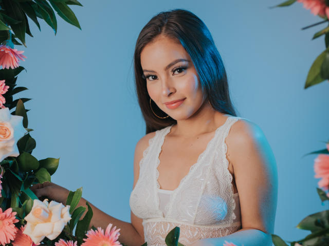 Kiaras Kopf und die oberen braunen Haare sind über eine graue Mauer sichtbar, mit rosa Blumen links und rechts, vor einem blauen Himmel. Ihr Gesicht ist größtenteils verdeckt..
