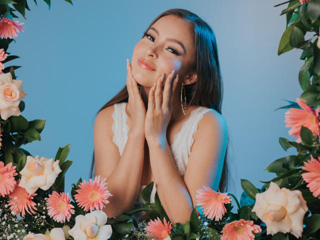 KiaraFire, con lunghi capelli castani, sorridente, mani sulle guance, indossa una camicetta bianca in pizzo, circondata da fiori rosa e bianchi, luce blu, orecchini dorati, dall'aspetto carina e sensuale..