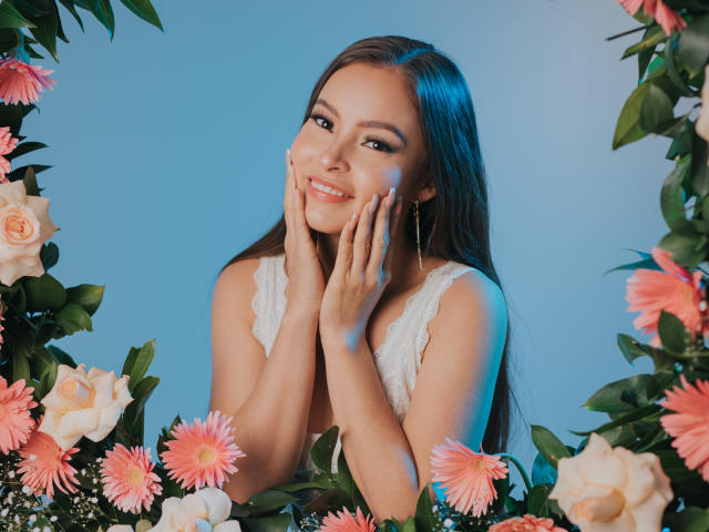 KiaraFire with long brown hair, smiling, hands on cheeks, wearing white lace dress, surrounded by pink and white flowers, blue lighting. Boobs, cute, pretty.