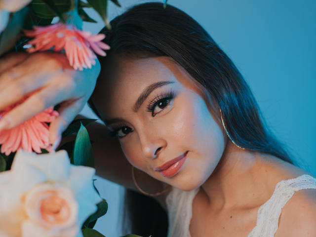 KiaraFire with long dark hair, hoop earrings, and glossy pink lips, looking seductively at the camera, surrounded by pink and white flowers, in a blue-lit setting.