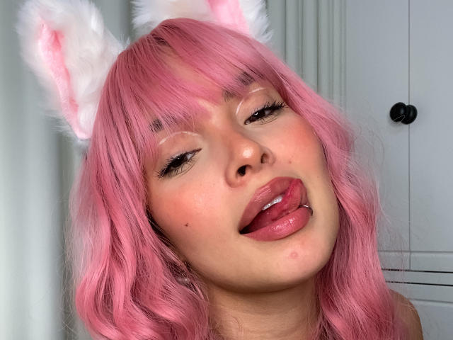 KiaraFire, con i capelli rosa, orecchie da gatto bianche e soffici, e la lingua leggermente fuori, guarda in alto con gli occhi socchiusi, mostrando un piccolo brufolo sulla guancia..