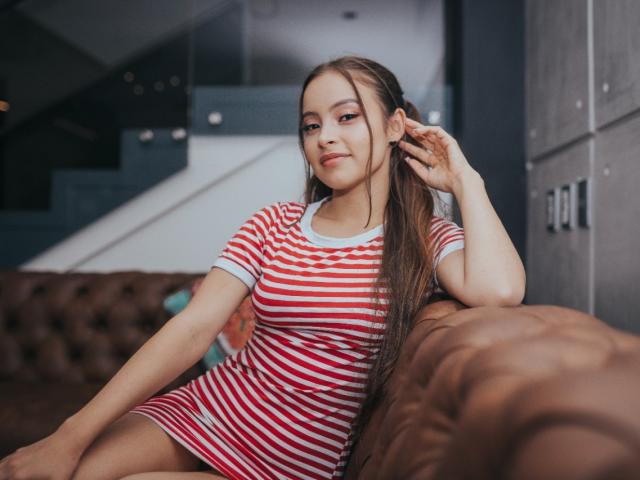 KiaraFire with long brown hair, red and white striped dress, sitting on brown tufted sofa, hand in hair, looking slightly to the side, subtle smile, cute, small tits.