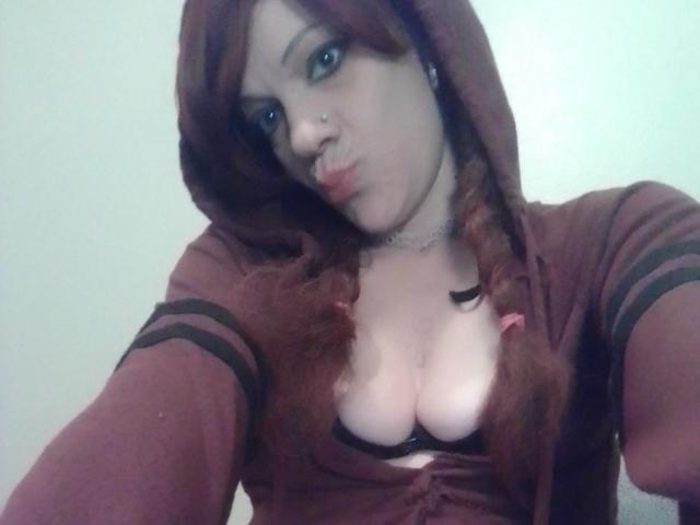 FreakySaint com cabelo ruivo em tranças, lábios carnudos, vestindo um moletom vinho, mostrando seios grandes em um sutiã preto, com um piercing no nariz e um colar, tirando uma selfie..