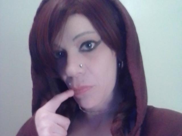 FreakySaint, con cabello rojo, piercings en la nariz y delineador negro, chupándose el dedo, vistiendo una sudadera marrón. Tiene una expresión linda, ligeramente arrogante..