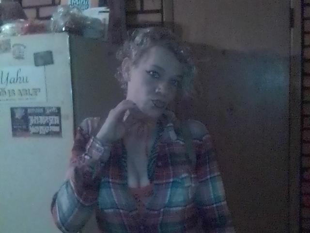 FreakySaint has curly blonde hair, wears a red and blue plaid shirt with a low neckline, showing tits, and stands in front of a slightly fogged window.