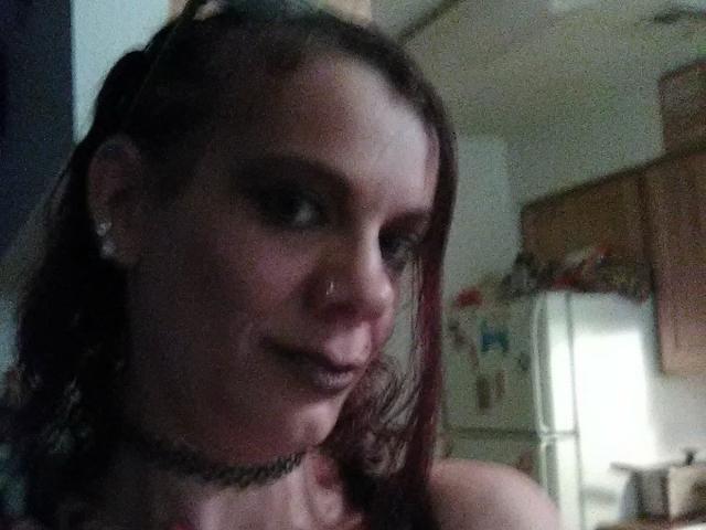 FreakySaint tiene el cabello castaño oscuro, un collar, y un piercing en la nariz, con maquillaje de ojos ahumado y una ligera sonrisa, en una habitación con poca luz. Sus senos son parcialmente visibles..