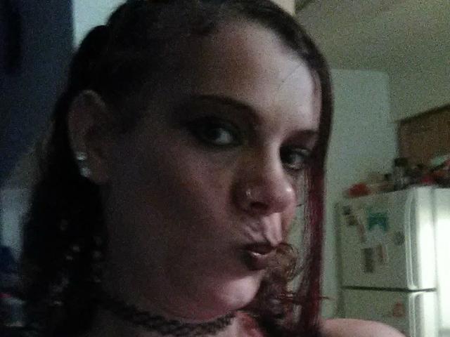 A FreakySaint tem lábios volumosos, maquiagem escura nos olhos e um piercing no nariz. Ela está usando um colar e tem cabelo trançado vermelho, com uma expressão séria. Seus seios estão parcialmente visíveis..