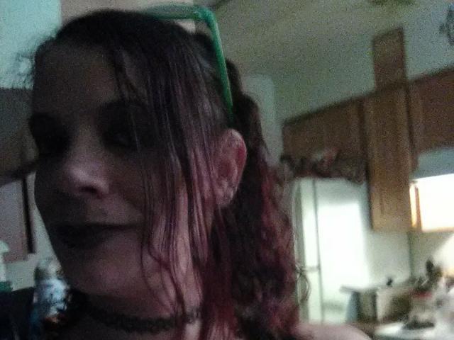 FreakySaint com maquiagem escura, faixa de cabeça verde e cabelo tingido de vermelho, usando batom preto e colar, em uma cozinha com pouca luz. Seios não visíveis, foco no rosto..