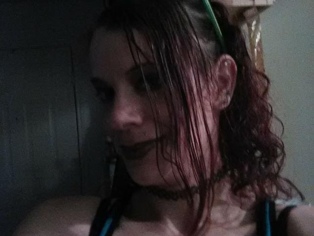 FreakySaint has wet brown hair, green headband, black choker, and dark lipstick, with a slightly blurred, darkly lit face showing a subtle smile.
