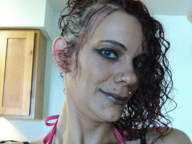 FreakySaint hat lockiges, rotes Haar, blaue Augen und ein Nasenpiercing mit einem kleinen Stecker. Sie trägt dunkelroten Lippenstift und ein pinkes Halstuch-Top und lächelt leicht..