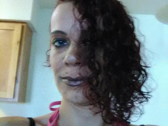 FreakySaint con i capelli rossi ricci che le coprono un occhio, ombretto blu, piercing al naso, rossetto viola lucido, indossa un top rosa a fascia, in un selfie leggermente sfocato..