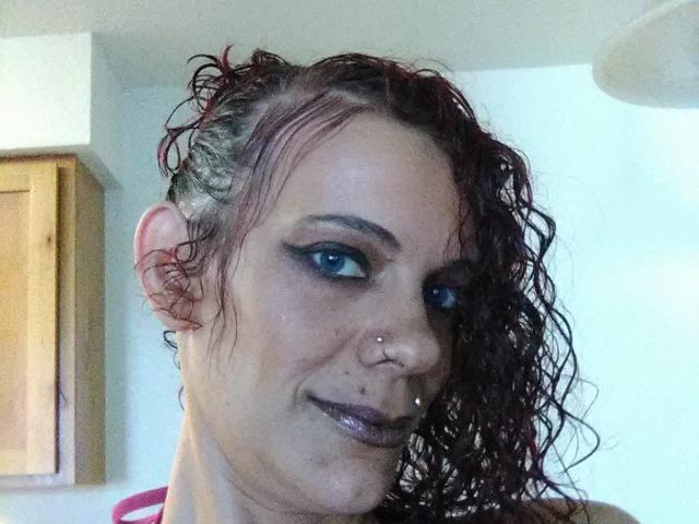 FreakySaint heeft nat, krullend rood haar, blauwe oogmake-up, neusringen en een lipring, en ze glimlacht met een licht rommelig gezicht. Haar borsten zijn niet zichtbaar..