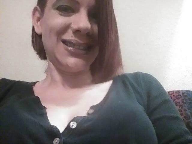 FreakySaint, com cabelo castanho curto, batom preto e um sorriso, vestindo uma camisa preta de botões que mostra seus seios, em frente a uma parede lisa. Ela está olhando para a câmera..