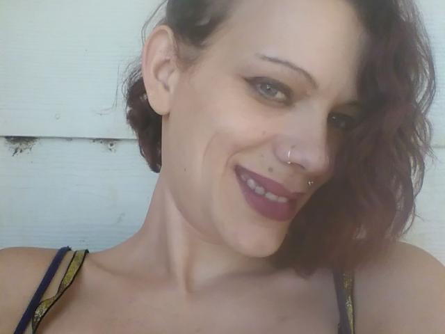 FreakySaint, avec ses cheveux bruns ondulés, son anneau de nez et son rouge à lèvres foncé, sourit à la caméra, montrant ses seins dans un haut coloré. Sa peau est claire et elle a l'air adorable..