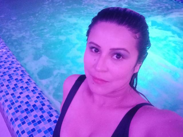 Contelaya a les cheveux bruns et mouillés, elle porte une combinaison noire, et on peut voir ses seins dans une piscine éclairée en bleu. Elle sourit légèrement, et il y a un bord carrelé sur la gauche..