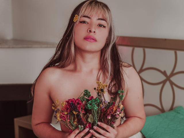 SallyDoll con capelli biondi e castani, rossetto rosa, che tiene un bouquet di fiori colorati, coprendosi il seno, che guarda verso la fotocamera, con una molletta gialla tra i capelli, e uno sfondo con un cuscino verde..