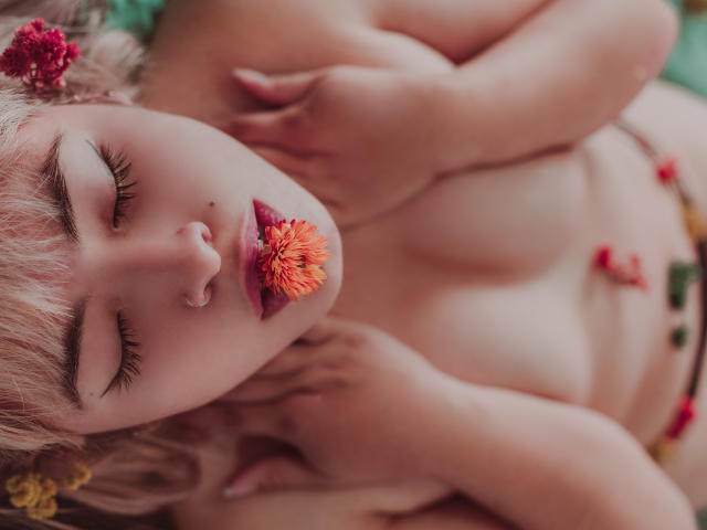 SallyDoll con capelli rosa, occhi chiusi, un fiore in bocca, nuda, mani sul petto, piccoli seni, pelle pallida, naso rosa, copricapo floreale e petali di fiori colorati sul corpo..
