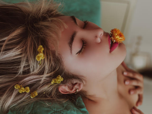 SallyDoll con capelli biondi, fiori gialli tra i capelli, occhi chiusi, fiore arancione in bocca, rossetto rosa, piccolo anello al naso, mani sul petto, sfondo verde, illuminazione soffusa, sensuale..