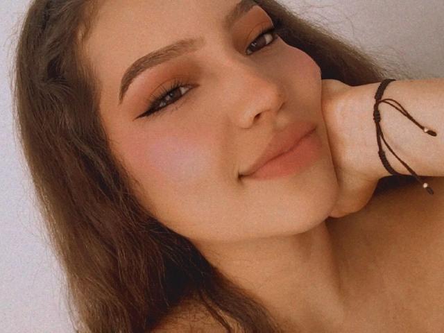 Tammy Andrade mit langem, braunem Haar, dicken Augenbrauen und einem leichten Lächeln, die ihr Kinn in die Hand gestützt, zeigt ihre weiche, rosa geschwungene Vulva und ihren niedlichen Hintern..