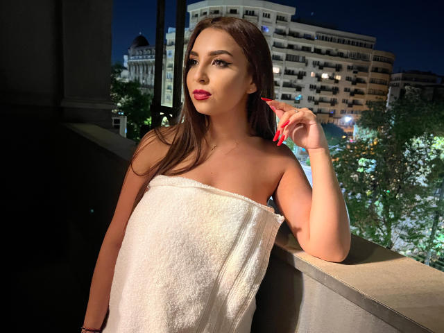 KayaOcean mit langem, braunem Haar, rotem Lippenstift und roten Nägeln, trägt ein weißes Handtuch, lehnt sich an einen Balkon, hält ihr Haar und im Hintergrund sind die Lichter der Stadt bei Nacht zu sehen..