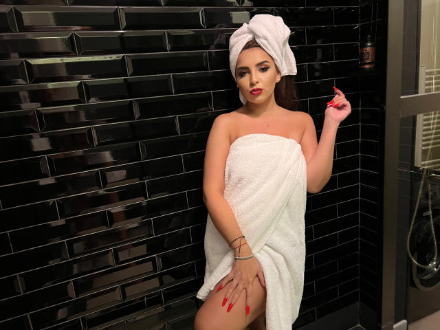 KayaOcean with a white towel wrapped around her pussy, black tiles in the background, red nails, towel on her head, hand on thigh, looking at camera.