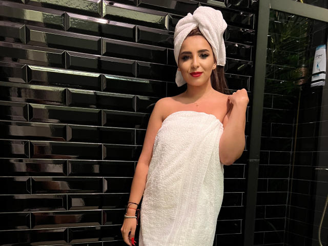 KayaOcean with a white towel wrapped around her tits and ass, dark brown hair in a towel, black tiled background, red lipstick, black bracelets, and a slight smile.