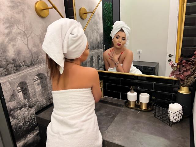 KayaOcean with white towel on her ass, looking in mirror, has towel on head, red lipstick, golden wall sconces, black tiles, and white toiletries on gray countertop.