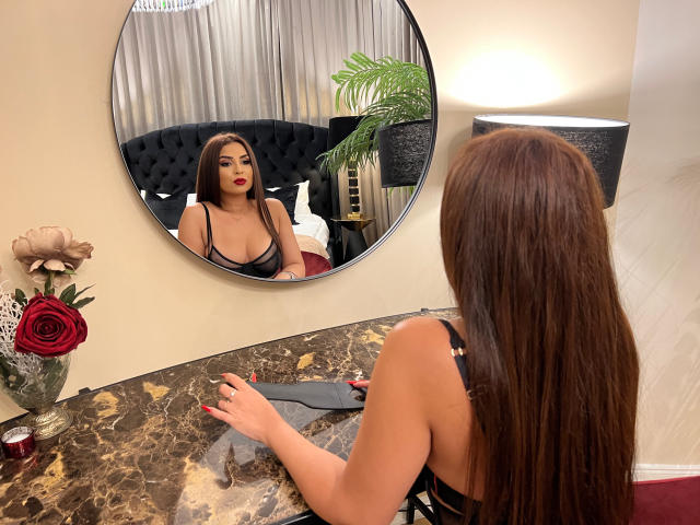 KayaOcean, com longos cabelos castanhos, vestindo lingerie preta, olha para um espelho, mostrando seus seios, batom vermelho e segurando um pano preto em uma bancada de mármore..