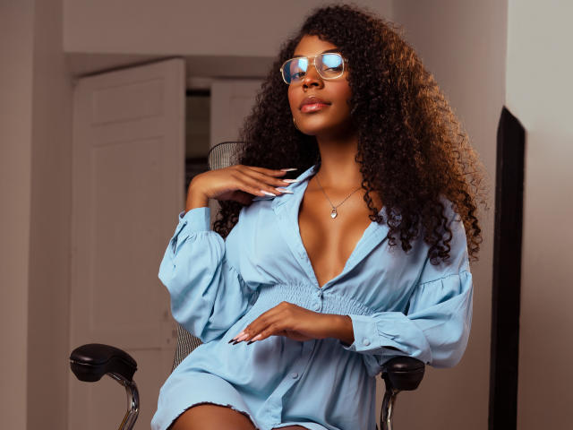 TianaParkerr, avec ses cheveux bruns bouclés, portant une robe bleue à boutons, des lunettes reflétant la lumière, assise sur une chaise, une main sur sa poitrine, montrant ses seins, dans une pose détendue..