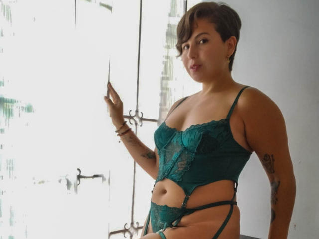 Alice Howards, com cabelo castanho curto, vestindo um conjunto de lingerie verde de renda com recortes, em pé perto de uma janela, mostrando seus seios, tatuagens nos braços e um olhar confiante e sexy..