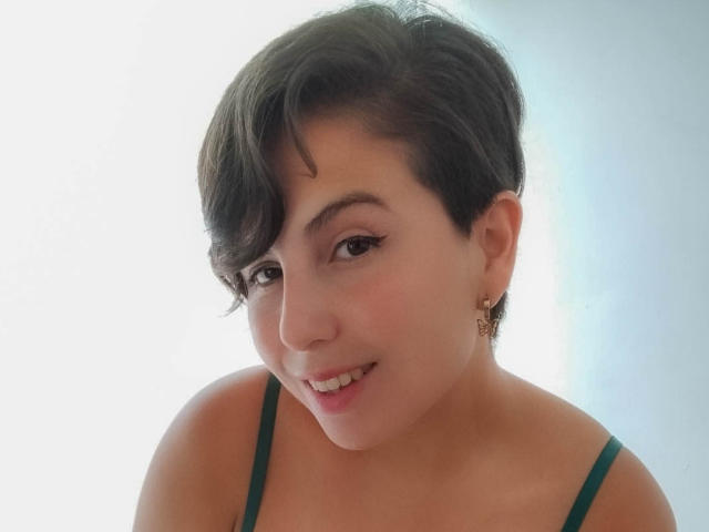 Alice Howards, con cabello negro corto, tirantes verdes, aretes pequeños, sonriendo, piel clara, ojos marrones, linda, labios rosados suaves, parte superior del cuerpo, ligera inclinación, mirando a la cámara, luz natural..