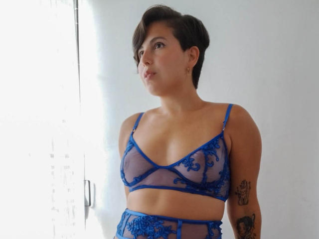 Alice Howard mit kurzen, braunen Haaren, trägt einen blauen Spitzen-BH und ein passendes Set aus Strümpfchen. Sie hat Tattoos auf ihrem rechten Arm und blickt zur Seite. Sie hat kleine Brüste..