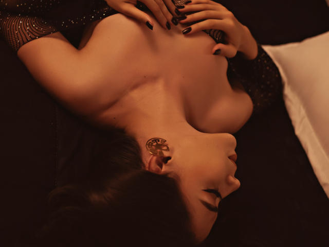 Mara Moretti, nue, allongée sur le dos, les mains sur ses seins, portant une boucle d'oreille dorée, du vernis à ongles noir et une manche noire pailletée, dans un décor sombre et peu éclairé..