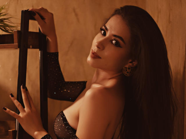 Mara Moretti, con cabello oscuro, vestida con una blusa negra brillante, sosteniendo un marco negro, mirando por encima del hombro con una sonrisa seductora, mostrando sus hombros desnudos y sus aretes..