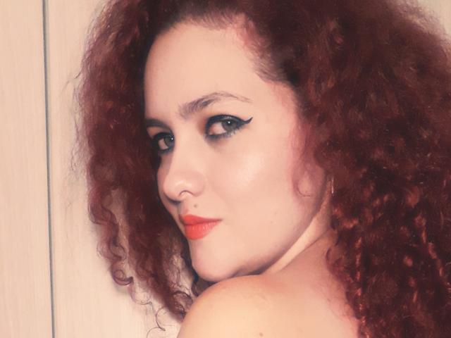 GinebraNova ha i capelli rossi ricci, l'eyeliner nero ad ali di farfalla e un rossetto rosso acceso, e guarda oltre la spalla con un sorriso appena accennato. Le sue spalle scoperte sono visibili..
