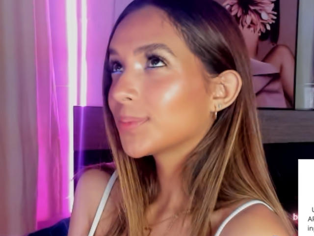 Sarah West, con la pelle luminosa, i capelli castani e un sorriso discreto, indossa una maglietta bianca, piccoli orecchini e guarda di lato. Il suo seno è piccolo e i suoi glutei non sono visibili..