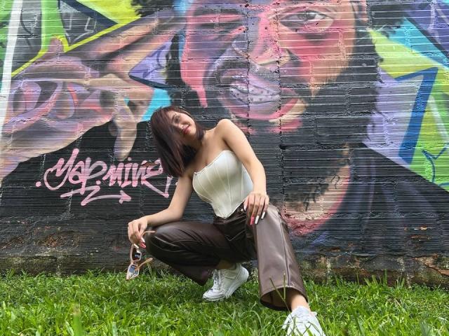 EstefaniaRamos: In einer tiefen Hocke, trägt ein weißes Top ohne Träger, braune Hosen und weiße Turnschuhe, mit kurzen schwarzen Haaren, vor einer bunten Graffiti-Wand auf grünem Gras..