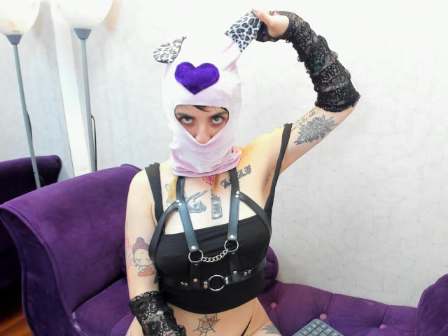 AmySpider, con tatuaggi, indossa una maschera bianca con un cuore viola, un top corto nero con anelli metallici, guanti di pizzo nero e orecchie da gatto bianche, seduta su un divano viola..