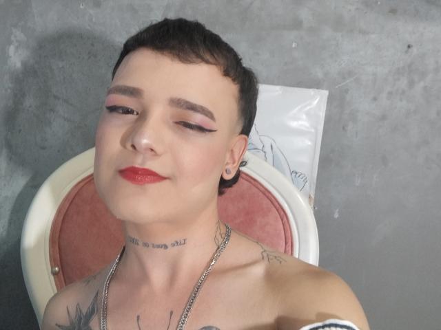 HeidySumers with short brown hair, red lipstick, and pink eyeliner, smiling, wearing a chain necklace, tattoos on neck and chest, seated against a red and white chair.