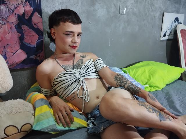 HeidySumers with short black hair, red lipstick, striped off-shoulder top, denim shorts, tattoos, and small tits, sitting on bed with green pillow, striped blanket, and stuffed toy.