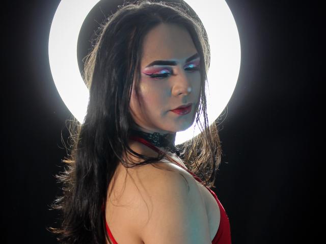 Iris Durand, con el pelo oscuro y mojado, un vestido rojo y maquillaje de ojos colorido, de perfil, con una luz circular blanca brillante detrás, un collar negro, lápiz labial rojo, pechos medianos, expresión seria, fotografía..