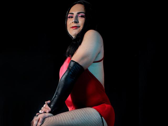 IrisDurand with long black hair, red makeup, red one-piece, black gloves, and fishnet legs, sitting, smiling, showing her red ass, and her white-painted nails against a black backdrop.