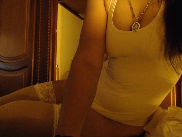 AfroditaHan in a tight white tank top, showing her tits and wearing white lace thigh-highs, with a necklace and a slightly visible pussy, in dim, yellow lighting.