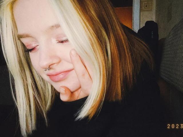 LaPochka, con capelli biondi e castani, piercing al naso, ombretto rosa e una mano sul viso, sorride dolcemente, indossando una maglietta nera, in una stanza poco illuminata.. 2022.