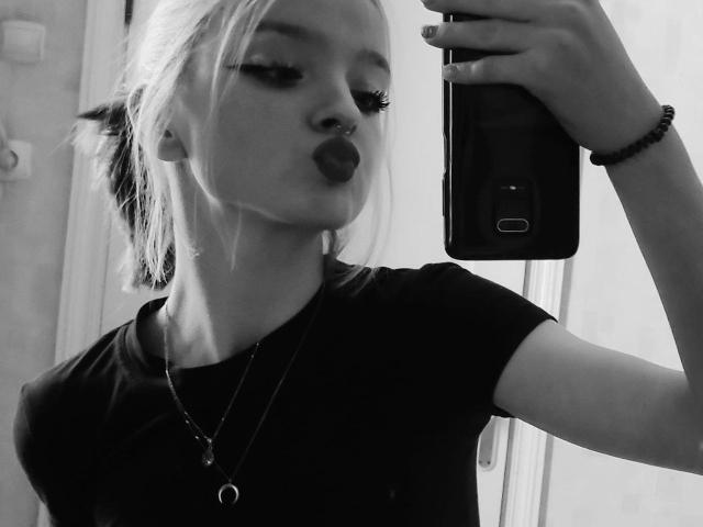 Selfie en blanco y negro de LaPochka con los labios fruncidos, un piercing en la nariz, pestañas largas, una camiseta negra, un collar y una pulsera, sosteniendo un teléfono, con su cabello rubio recogido en un moño despeinado..