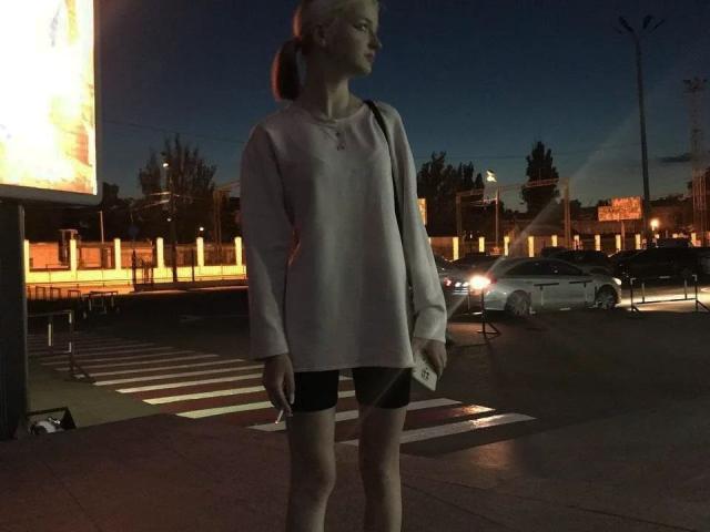 LaPochka stands at a crosswalk, wearing a light pink sweater and black shorts, holding a phone, with a car's headlights in the background, and a dark evening sky.
