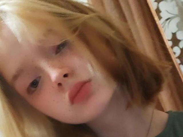 LaPochka, avec ses cheveux blonds légèrement en désordre et ses taches de rousseur, ses lèvres rosées et une expression neutre, portant une chemise verte, sur une photo de profil floue..