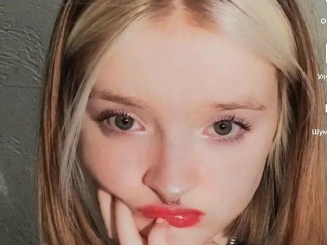 O rosto de LaPochka, mostrando cabelo loiro, olhos verdes, lábios vermelhos, um piercing no nariz, e a mão tocando o queixo, com o traseiro ligeiramente visível na parte inferior..