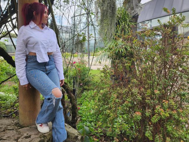NicoColeman with red hair, white button-up shirt tied at the front, ripped blue jeans, and white sneakers, leaning against a wooden pole in a lush green garden.
