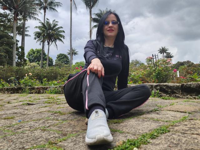 AlanaMoneth with black hair, blue sunglasses, and black outfit, sitting on stone path with white sneakers, palm trees in background, cloudy sky, and green plants, looking at camera.